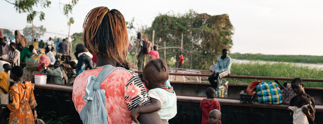 Eine Frau mit Baby auf dem Arm sieht Leuten zu, wie sie ein Boot einräumen. Viele Menschen im Südsudan sind auf der Flucht.