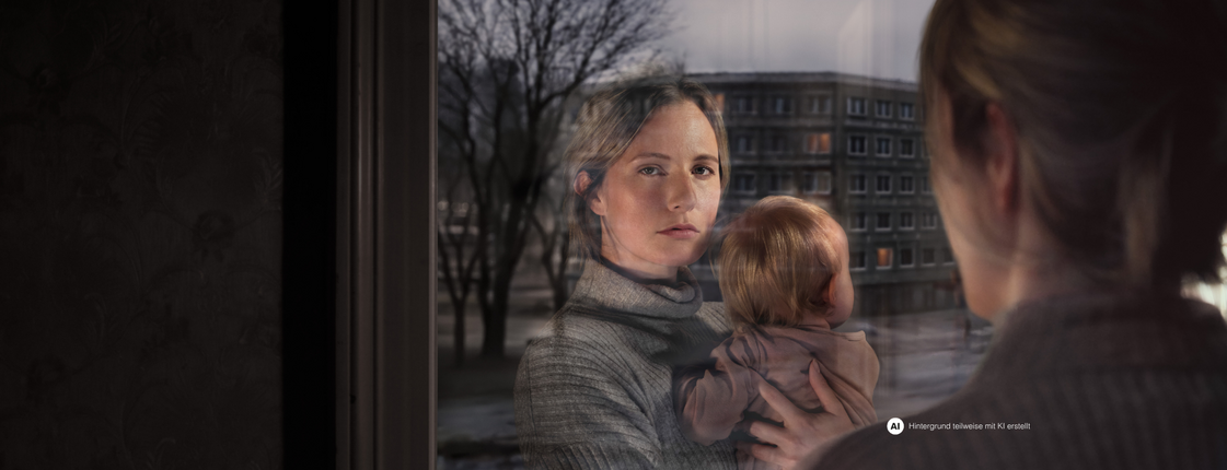 Eine Frau mit Kind am Arm schaut aus dem Fenster. Ihr Gesicht spiegelt sich im Fensterglas wider. Vor dem Fenster ist eine Wohngegend erkennbar.