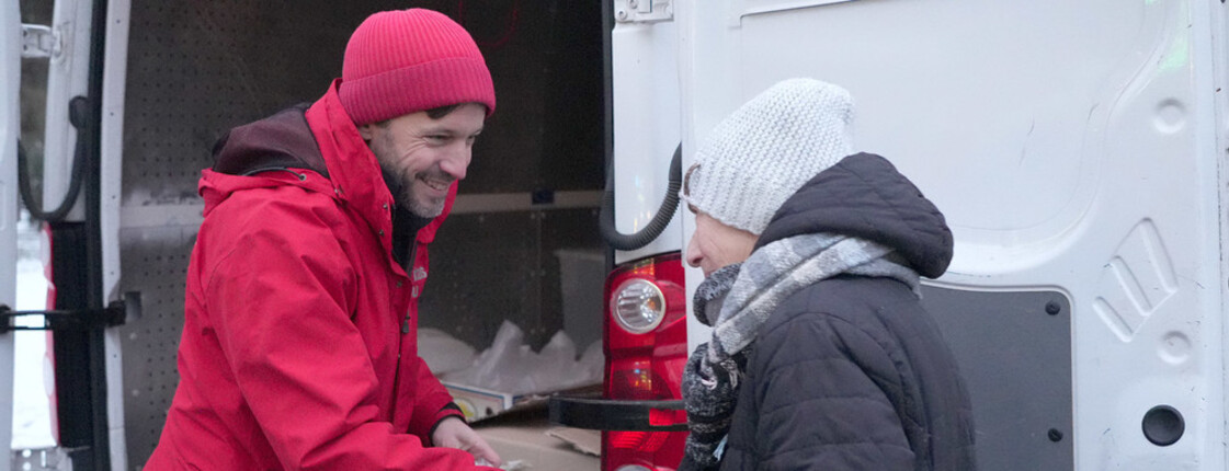 Ein Caritas-Mitarbeiter schüttelt einer winterlich gekleideten alten Dame die Hand und überreicht ihr Hilfsgüter. Im Hintergrund ist ein Transporter mit Kartons zu erkennen, in denen sich weitere Hilfsgüter befinden.