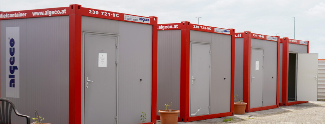 Caritas ZufluchtsDorf Mehrere rot-graue Container im Freien, gegenüber ein überdachter Sitzbereich.