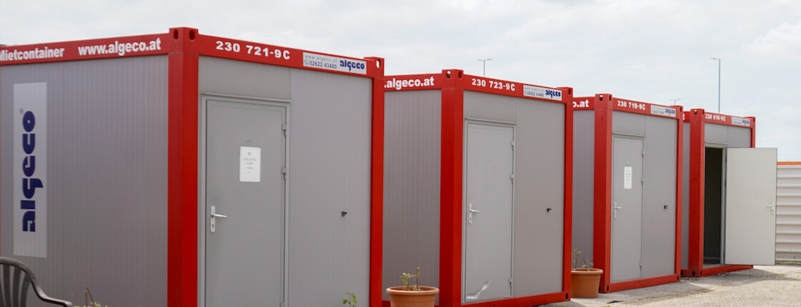 Mehrere rot-graue Container im Freien, gegenüber ein überdachter Sitzbereich.
