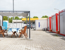 Bequeme Holzsessel und ein Holztisch mit Sonnenschutz. Im Hintergrund sieht man Container.