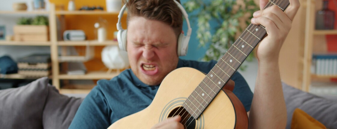Portrait eines Mannes auf einer Wohnzimmercouch, der weiße Kopfhörer trägt und leidenschaftlich akustische Gitarre spielt.