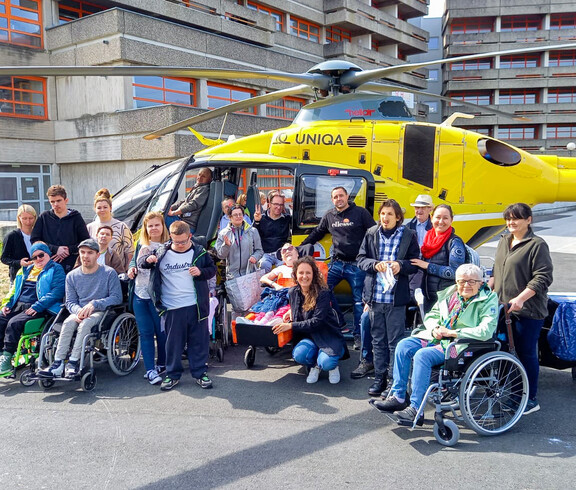 Bewohner*innen und Betreuer*innen des Haus St. Stephan waren am Tag der offenen Klinktür dabei. Sie stehen als Gruppe gemeinsam vor einem gelben Hubschrauber und lächeln in die Kamera.