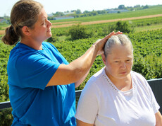 Eine Betreuerin in einem blauen Shirt kämmt einer Bewohnerin aus dem Haus Lisa draußen die Haare, während sie vor einer grünen Landschaft mit Feldern im Hintergrund stehen.