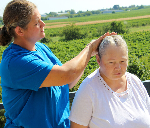 Eine Betreuerin in einem blauen Shirt kämmt einer Bewohnerin aus dem Haus Lisa draußen die Haare, während sie vor einer grünen Landschaft mit Feldern im Hintergrund stehen.