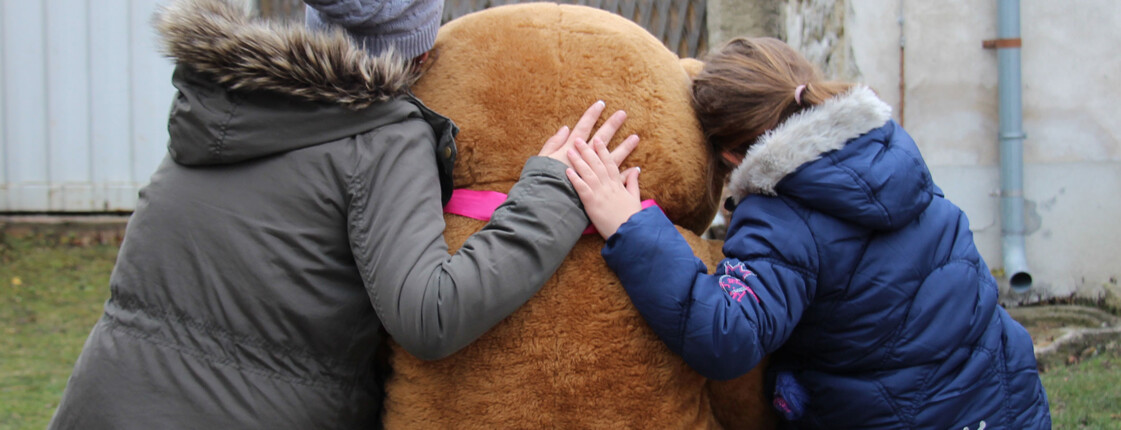 Eine Mutter sitzt mir ihrer Tochter und einem großen Teddy auf einer Mauer vorm Caritas Mutter+Kind=Haus.