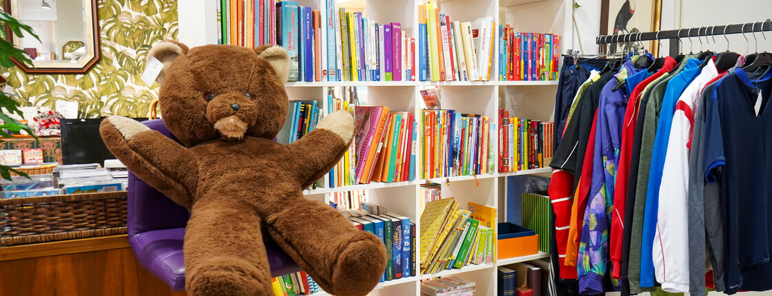 Ein Bereich des carla Shops im Burgenland mit einem großen braunen Plüschbären, einem Regal voller bunter Bücher und einem Kleiderständer mit Secondhand-Kleidung.
