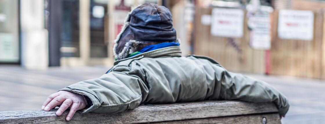 Eine Person in dicker Jacke und Mütze sitzt mit ausgestreckten Armen auf einer Holzbank, mit dem Rücken zur Kamera.