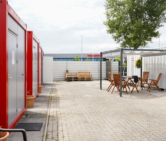 Mehrere rot-graue Container im Freien, gegenüber ein überdachter Sitzbereich.
