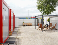 Mehrere rot-graue Container im Freien, gegenüber ein überdachter Sitzbereich.