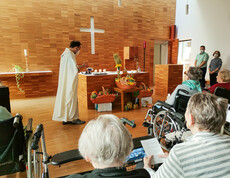 eine Erntedankmesse in einer Kirche. Ein Priester steht am Altar und hält den Gottesdienst, während Gemeindemitglieder, darunter ältere Menschen, in den Bänken sitzen und dem Geschehen folgen. Die Erntegaben sind festlich vor dem Altar arrangiert, und die Atmosphäre ist feierlich und besinnlich.