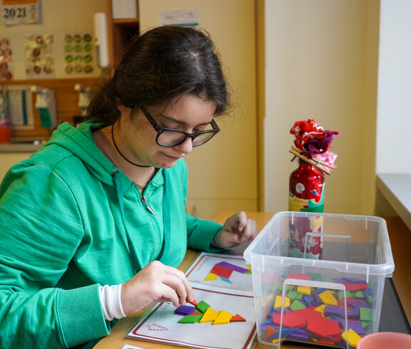 Eine junge Frau sitz an einem Tisch und macht ein Puzzle mit bunten Steinen.