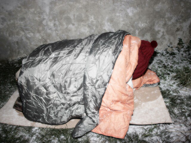 Obdachloser Mensch liegt in Schlafsack eingewickelt im Schnee auf einem Karton