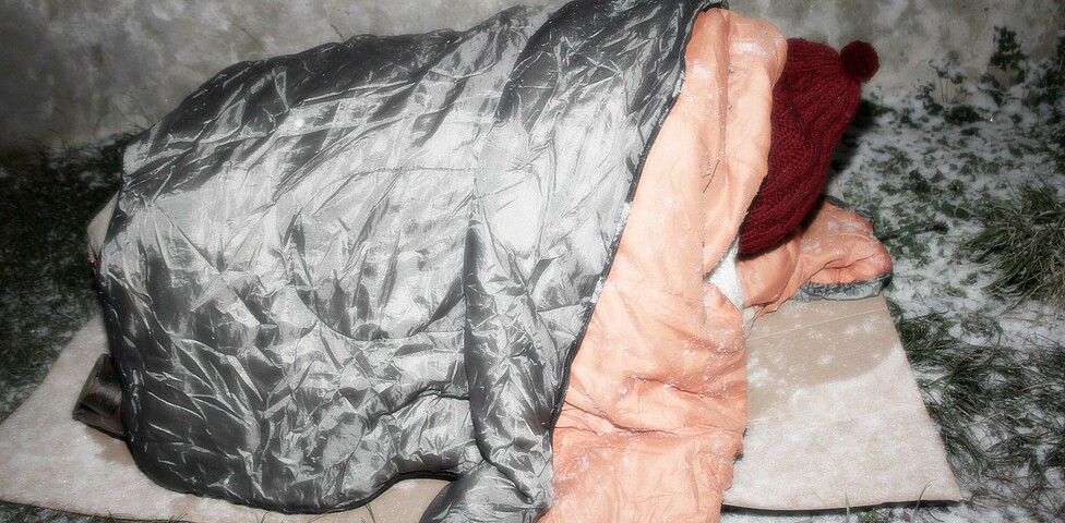 Obdachloser Mensch liegt in Schlafsack eingewickelt im Schnee auf einem Karton
