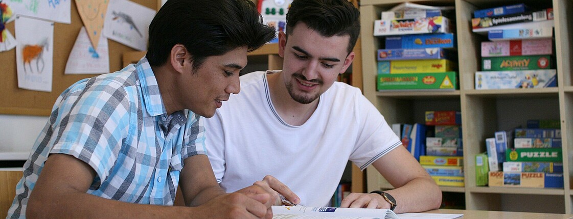 Zwei junge Männer sitzen an einem Tisch und schauen in ein Lernhilfebuch.
